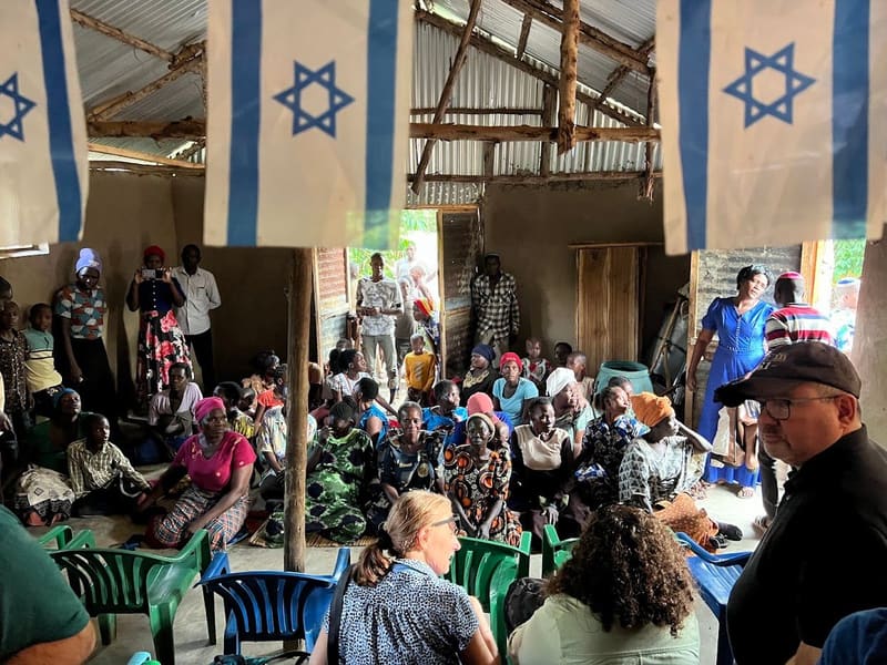 A packed community gathering in an open-sided hall hung with Israeli flags, with visitors seated in the foreground.