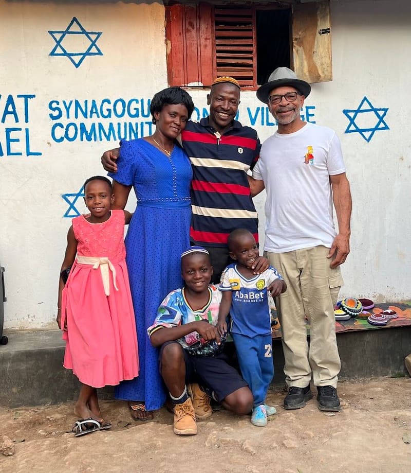 A family and a visitor pose in front of a wall painted "Ahavat Yisrael Synagogue Community Village" with blue Stars of David.