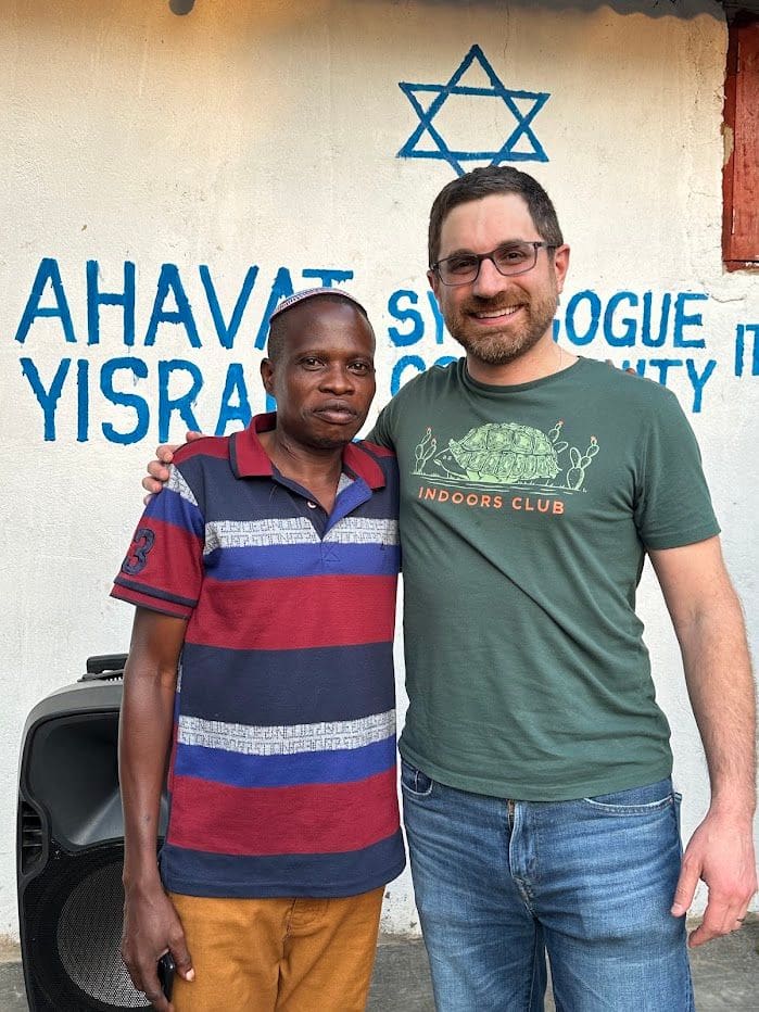 Two men with their arms around each other in front of the painted wall of the Ahavat Yisrael Synagogue Community.