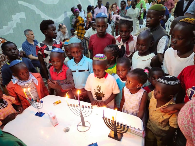 Children, several wearing kippot, crowd around a table to light Hanukkah menorahs at night.