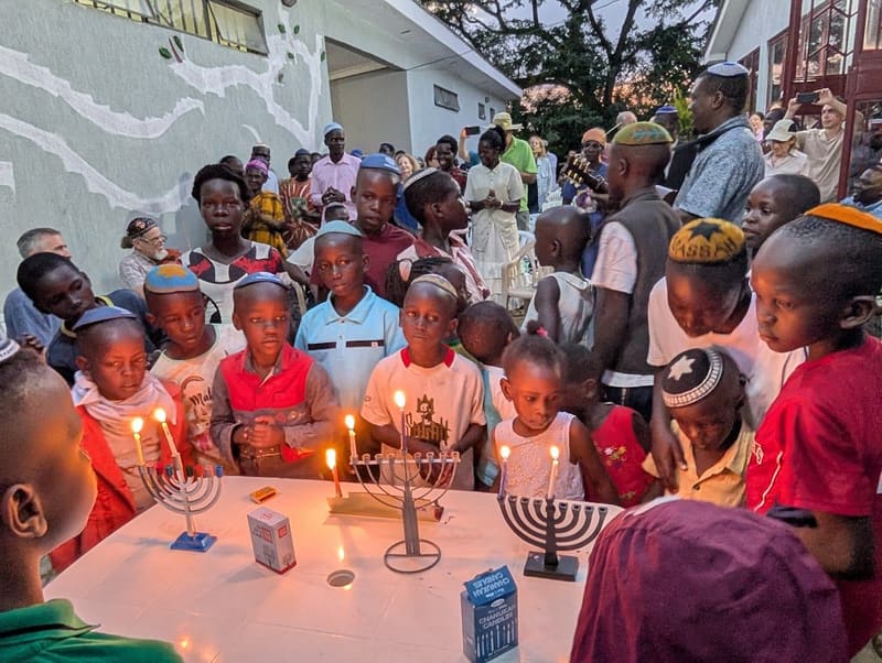 Children light Hanukkah candles on a table while a crowd of adults looks on.