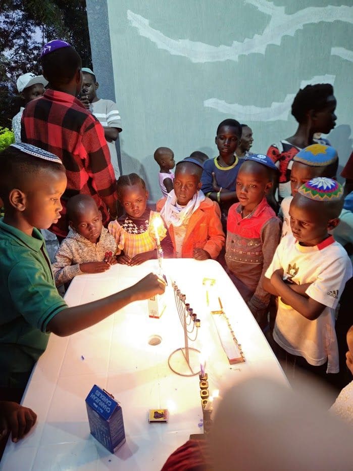 A boy lights a Hanukkah menorah on a white table while children, several wearing colorful kippot, watch in the candlelight.