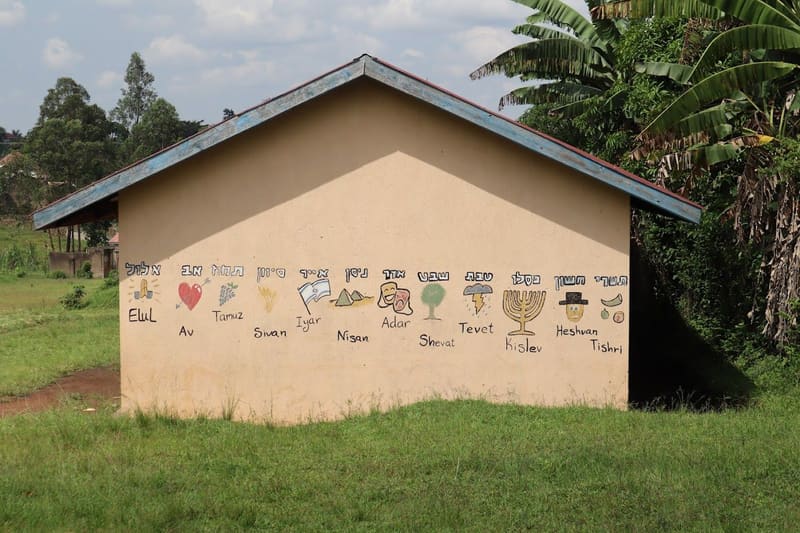 A school building painted with the months of the Hebrew calendar, from Tishri to Elul, each with a small illustration, beside banana trees.