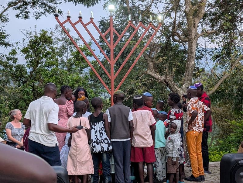 Adults and children gather beneath a large outdoor metal menorah at dusk.