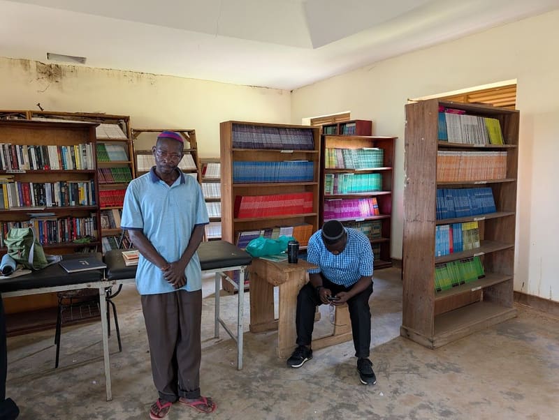 A school library lined with wooden bookshelves. One man in a kippah stands while another sits.