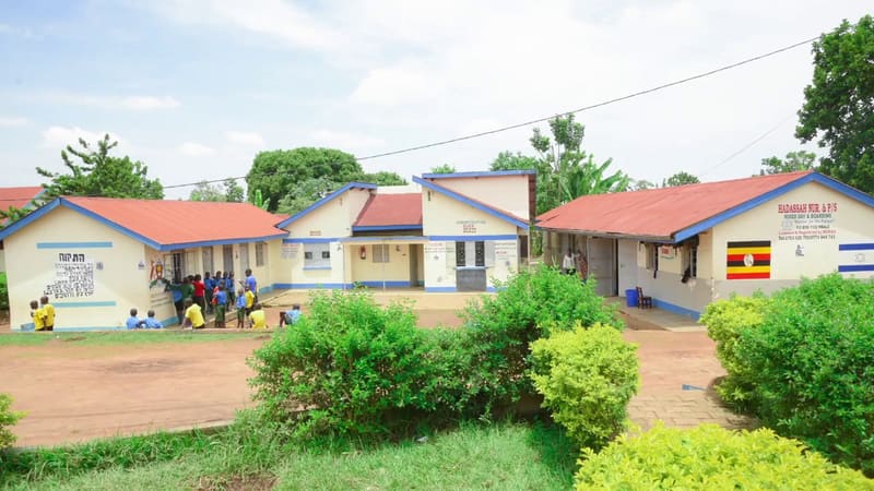 The Hadassah school campus: cream buildings with red roofs and blue trim, one painted with Hatikvah and another with the Ugandan flag, and children in the yard.