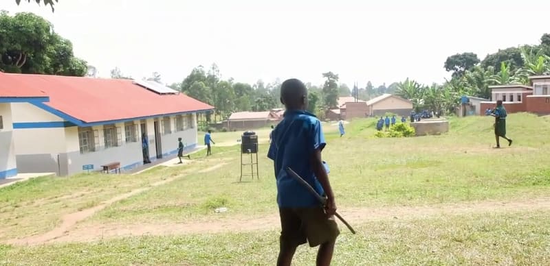 A Hadassah pupil in a blue uniform walks across the grassy campus towards children playing by the red-roofed classrooms.