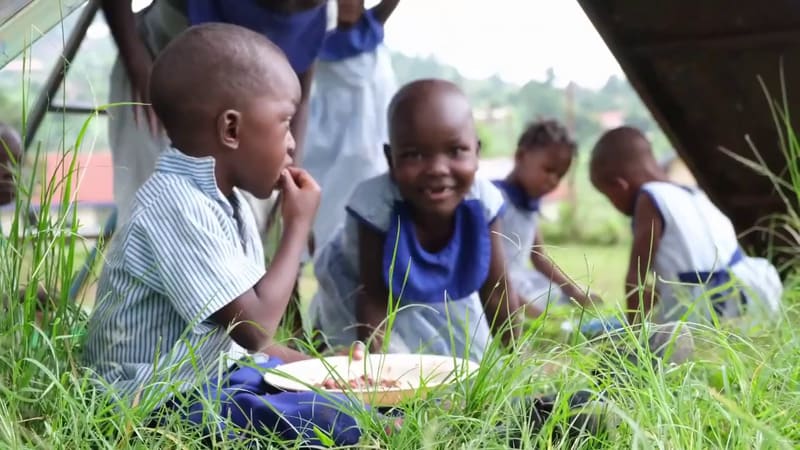 Young pupils at Hadassah eat their lunch sitting on the grass, one of them smiling at the camera.