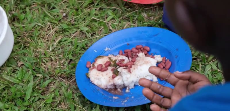 A blue plate of posho and beans on the grass at Hadassah, a child's hand reaching for it.