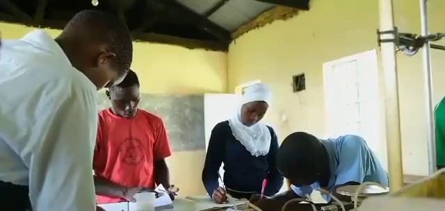 In the Semei Kakungulu High School science laboratory, a student in a white headscarf writes in a notebook beside classmates.