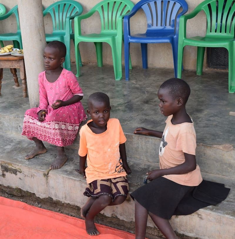 Three young children sit on a veranda step, with green and blue plastic chairs behind them.