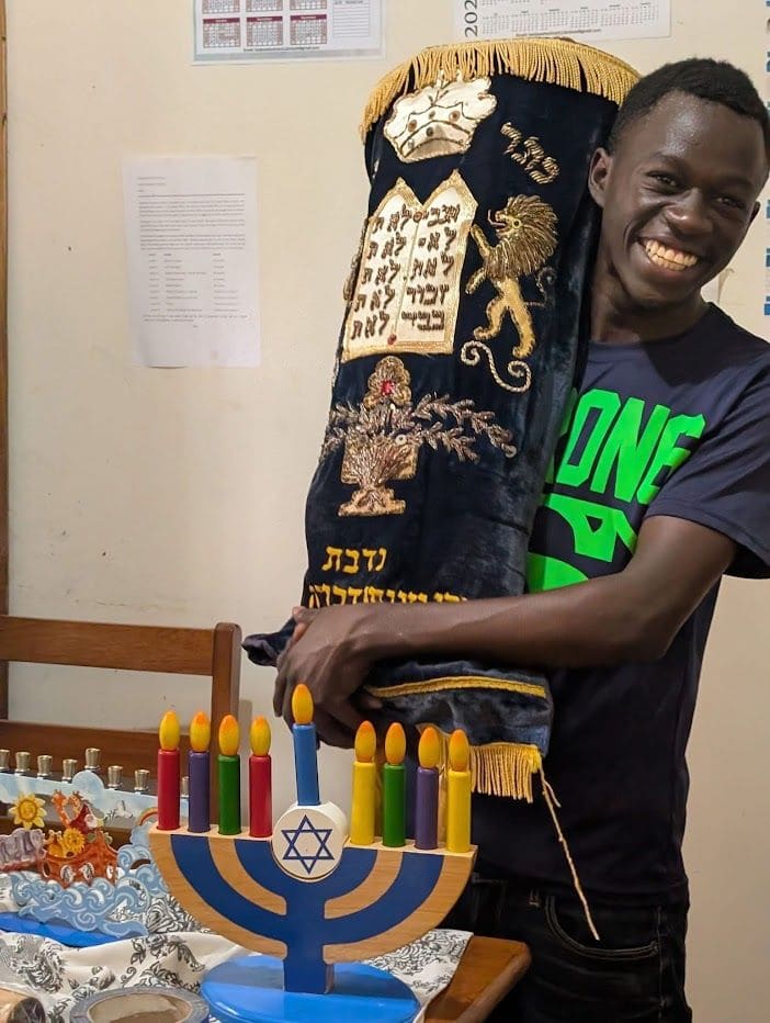 A beaming young man hugs a Torah scroll in an embroidered velvet cover. A colorful toy menorah stands in front of him.